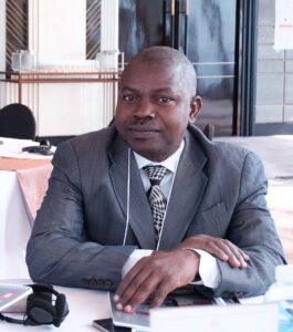 Image of Ganiyu Salau sitting at a table, looking directly at the camera. His hands are folded in front of him. He wars a gray suit, a white shirt and a patterned tie of silver and blue.