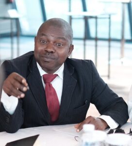 Photo of Ganiyu Salau. He is wearing a black suit with a white shirt and thick red tie. He is in the middle of talking and pointing at something with a pen.
